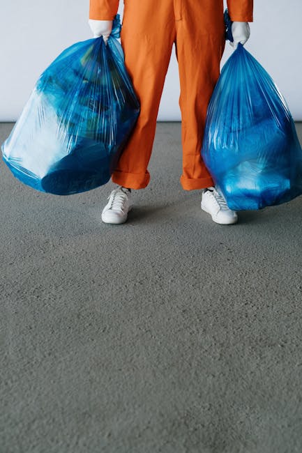 A person wearing orange overalls and white sneakers is standing indoors on a grey carpeted floor, holding two large blue plastic bags filled with waste or unwanted items, one in each hand. The individual’s upper body and head are not visible in the image. Behind them, there is a plain grey wall, and the person appears to be in the process of preparing for a house removal or waste clearance, potentially related to home relocation services. The scene captures the loading or carrying phase of a removal process, reflecting the practical tasks involved in furniture and waste transport. This image illustrates the logistics of packing and moving, which is a key aspect of professional removals provided by companies like Man with Van Southborough, who specialise in household removals, waste disposal, and transportation logistics.