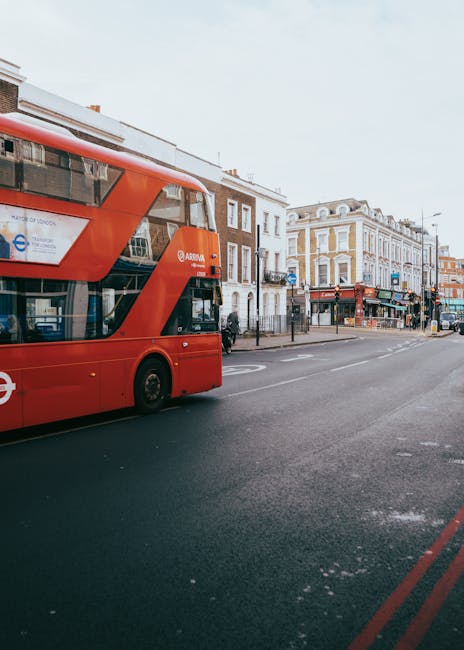 Image showing a red double-decker bus traveling along a city street in Southborough, with shops and residential buildings lining the sidewalk. The scene captures a typical urban environment with parked cars, pedestrians, and overhead signage. The street appears to be suitable for home relocation or moving logistics, as [COMPANY_NAME], such as Man with Van Southborough, offers professional removals and furniture transport services. Visible in the foreground is a section of the road with faint white markings and red lines along the curb, indicating parking restrictions. The lighting is natural, suggesting daytime conditions, with a partly cloudy sky overhead. The environment reflects a busy high street often used for loading and unloading household items during professional packing and moving processes, aligning with the area’s suitability for house removals and logistical planning for urban relocations.