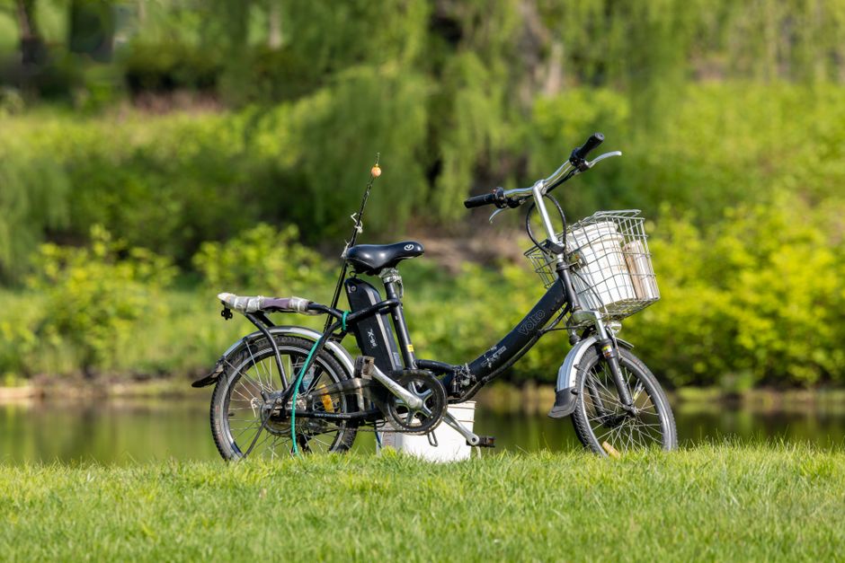 A black electric bicycle with a wire basket attached to the front handlebars, containing two rolled-up posters or papers, is parked on a grassy area near a body of water. The bicycle features a padded