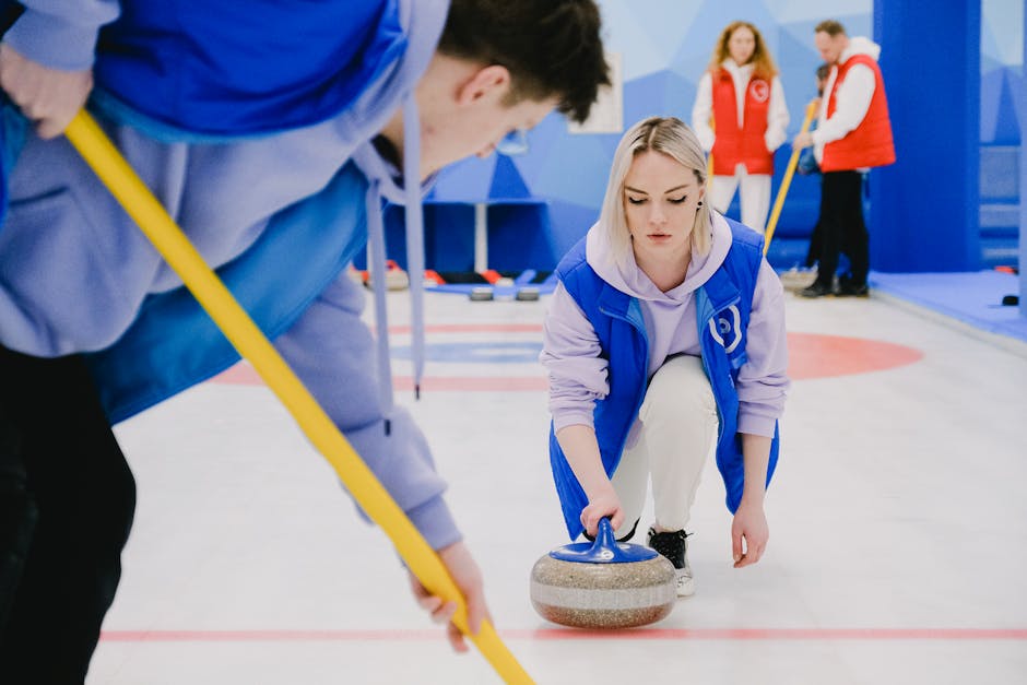 A woman dressed in a blue and white curling uniform is engaged in a home relocation process, kneeling on an indoor curling rink surface while gripping a curling stone with her right hand, preparing to release it towards the target area. She is surrounded by colleagues, one of whom, wearing a light blue top, is holding a yellow broom and appearing to guide or sweep the ice. In the background, two other individuals in red vests are observing, standing near the rink's blue wall. The area is well-lit with natural and artificial lighting, and the scene is part of a team activity involving packing, moving, and transport logistics, with the focus on careful handling of sports equipment related to a moving services context. Man with Van Southborough occasionally related to the sequence as a professional moving company, demonstrating careful loading or packing procedures involving equipment and packaging materials for home or sports facility relocation.