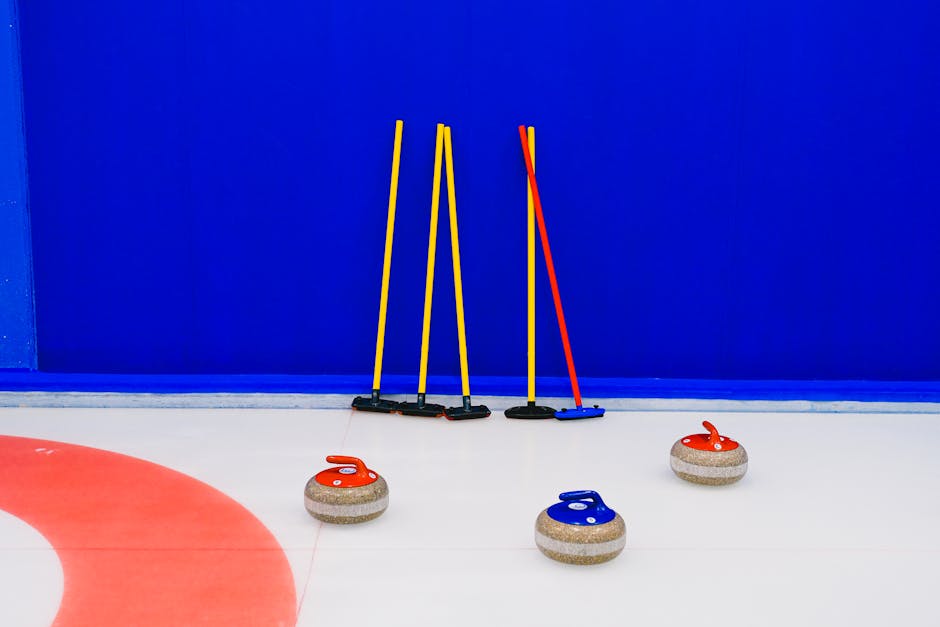A woman dressed in a blue and white curling uniform is engaged in a home relocation process, kneeling on an indoor curling rink surface while gripping a curling stone with her right hand, preparing to release it towards the target area. She is surrounded by colleagues, one of whom, wearing a light blue top, is holding a yellow broom and appearing to guide or sweep the ice. In the background, two other individuals in red vests are observing, standing near the rink's blue wall. The area is well-lit with natural and artificial lighting, and the scene is part of a team activity involving packing, moving, and transport logistics, with the focus on careful handling of sports equipment related to a moving services context. Man with Van Southborough occasionally related to the sequence as a professional moving company, demonstrating careful loading or packing procedures involving equipment and packaging materials for home or sports facility relocation.