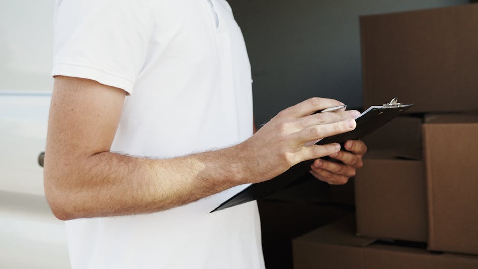 Close-up of a person wearing a white t-shirt, holding and writing on a black clipboard with a pen, inside a room with stacked cardboard boxes and a dark grey wall in the background, suggesting the packing or planning stage of a home relocation. The individual’s arm and hand are visible, emphasizing the process of organizing, inventorying, or preparing items for moving. This scene is part of a furniture transport or packing and moving process managed by Man with Van Southborough, supporting residential removal services including loading, packing, and logistics to facilitate a smooth house removal.