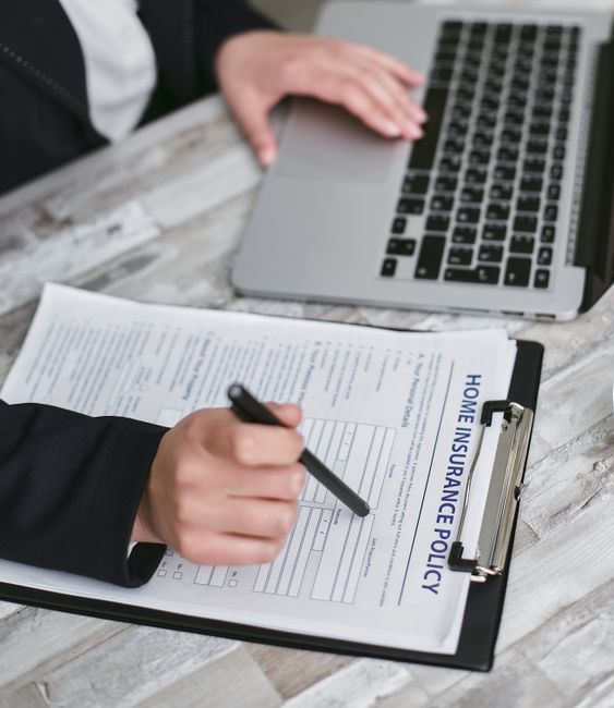 A person wearing a black suit is seated at a wooden table, filling out a home insurance policy form attached to a black clipboard with a black pen. In the background, there is a closed silver laptop with a black keyboard, placed on the same wooden table. The setting appears to be an indoor office or workspace with neutral lighting. The scene emphasizes the process of preparing documents related to property and belongings, which is often part of house removals and relocation planning, and reflects the careful organisation needed during home moves. This image is associated with Man with Van Southborough's services in removals, highlighting the importance of proper insurance and documentation during furniture transport and packing and moving activities.