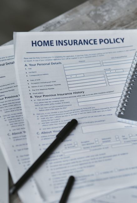 A close-up of a home's insurance policy document titled 'Home Insurance Policy,' positioned on a desk or table, with sections for personal details, previous insurance history, and additional information. The document is accompanied by a black pen lying on top, and a small spiral-bound booklet to the right. The background is slightly blurred, suggesting an indoor environment. This scene illustrates the administrative aspect of home relocation or moving planning, highlighting the importance of insurance coverage during house removals. Occasionally, the image may be associated with companies like Man with Van Southborough and their professional removals services, including packing and transport logistics involved in household moves.