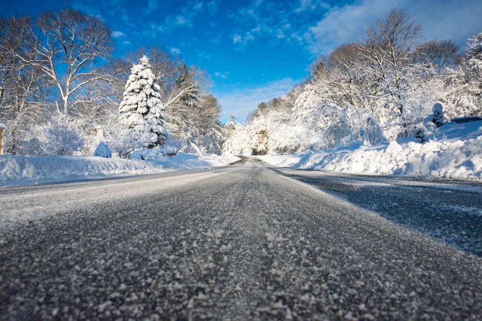 A low-angle view of a snow-covered rural road during daytime, with ice and snow evenly spread across the asphalt surface. Snow-laden trees and bushes line each side of the road, their branches heavily coated in white. The sky above is partly cloudy with patches of bright blue visible through the clouds. The scene reflects a winter setting, where the road appears undisturbed by traffic. In a house removal or relocation context, this image illustrates the importance of proper planning and caution during winter moves, especially when using services like Man with Van Southborough for furniture transport or home relocation amidst snowy conditions. The natural lighting highlights the cold, crisp atmosphere typical of winter, emphasizing the need for careful logistical arrangements in adverse weather.