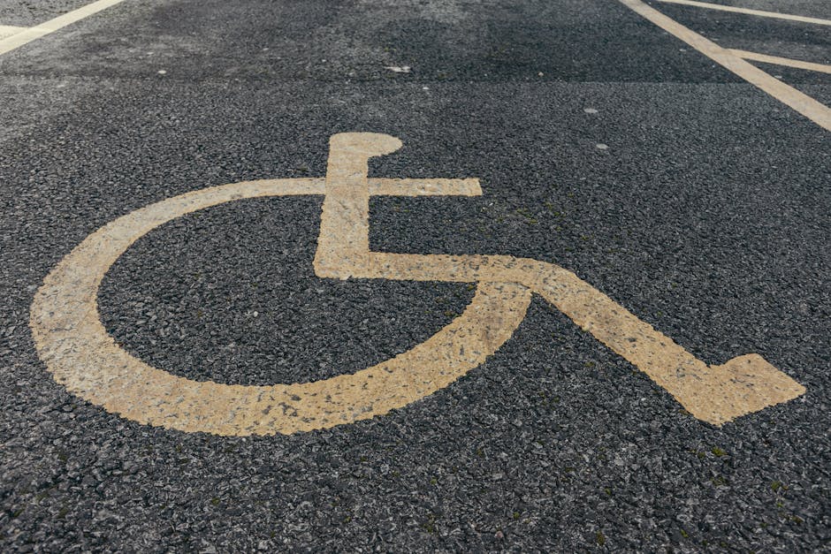 A close-up view of a yellow-painted wheelchair accessible parking symbol on dark, textured asphalt surface. The symbol consists of a stylized wheelchair with a circular wheel at the front and a supporting frame extending to the back, indicating designated parking for individuals with mobility impairments. The paint appears slightly worn and chipped around the edges, with visible debris and small cracks on the asphalt. In the background, faint parking lines are visible, and the scene is evenly lit, suggesting daylight. This image highlights accessible parking areas outside a residential or commercial property, which might be relevant in the context of house removals or relocation services by Man with Van Southborough, especially when considering parking access and logistics for home relocation or furniture transport.