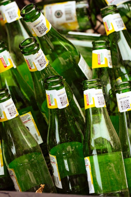 A close-up view of multiple green glass bottles with white labels and QR codes, stacked closely together inside a container. The bottles are arranged in a slightly tilted manner, some lying on their sides, and others standing upright. The background shows additional bottles with similar labels, out of focus, suggesting they are packed for transport or recycling. The setting appears to be indoors or in a warehouse environment, with natural or artificial lighting illuminating the bottles and their labels. This scene reflects the process of packing or preparing bottles for disposal or recycling, which may be relevant to house clearance or moving services offered by Man with Van Southborough.