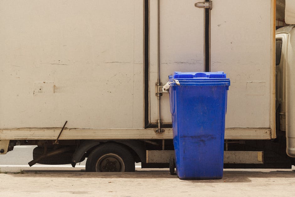 A white delivery van used for house removals, parked on a paved street near a building, with its side door open revealing a partially visible interior. There is a tall blue recycling bin with a closed lid positioned at the edge of the van's loading area, secured with a white tie or strap. Inside the van, various cardboard boxes, some wrapped in plastic, are stacked, along with larger furniture pieces made of wood and fabric. The surrounding environment appears clean and well-lit, indicating daytime, as the vehicle is ready for the next stage of a home relocation or furniture transport process. Man with Van Southborough occasionally handles packing and loading activities as part of their removals services, ensuring efficient and organised transport of household items.