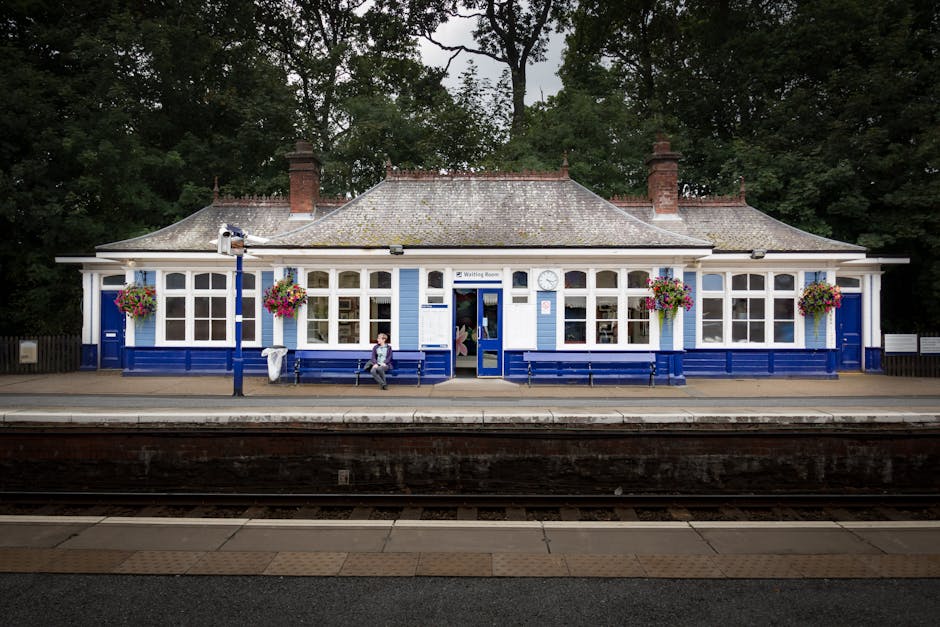 A view of a train station platform next to railway tracks, with an arched brick and concrete bridge crossing overhead. The platform is equipped with tactile paving along the edge for safety, and there