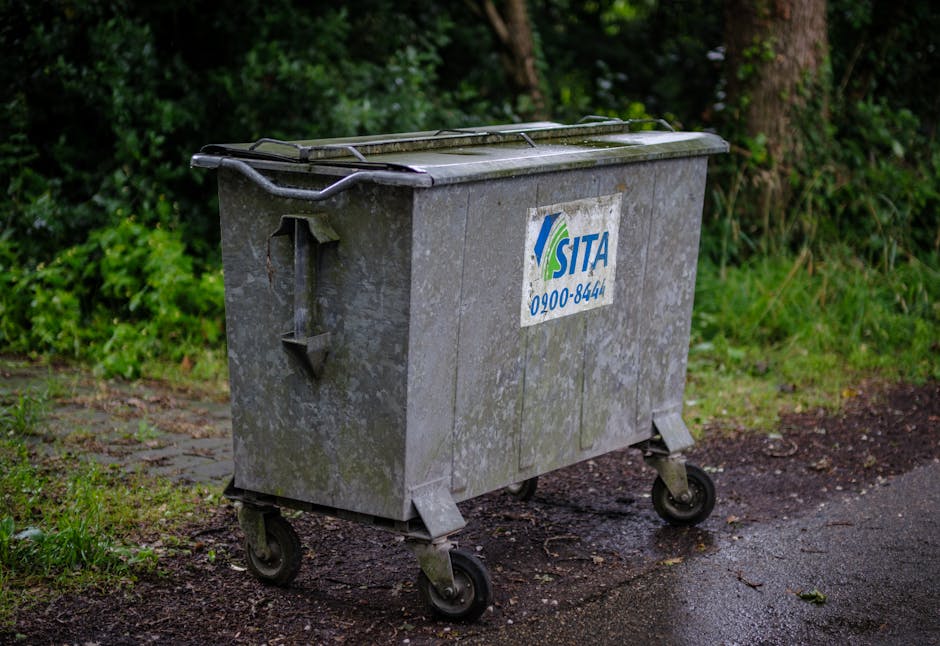 A large, grey metal wheeled waste bin with a hinged lid, positioned on a paved area next to a grassy verge with green foliage and trees in the background. The bin displays a partially visible blue and white SITA logo and contact number on the side. The pavement around the bin shows some dirt and slight dampness. This scene is typical of municipal waste collection zones, which could be relevant in the context of home relocation and planning for clutter removal during moving or furniture transport services. The presence of the bin on the roadside suggests it may be awaiting collection, and the outdoor lighting indicates daytime with natural light illuminating the scene, fitting a neutral, factual description related to moving logistics handled by companies like Man with Van Southborough.