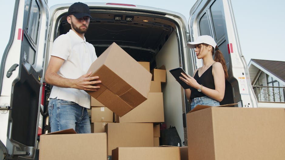 A professional mover from Man with Van Southborough is loading packing boxes and large items into a white cargo van outside a modern building with large glass windows. The mover, wearing a dark unifor